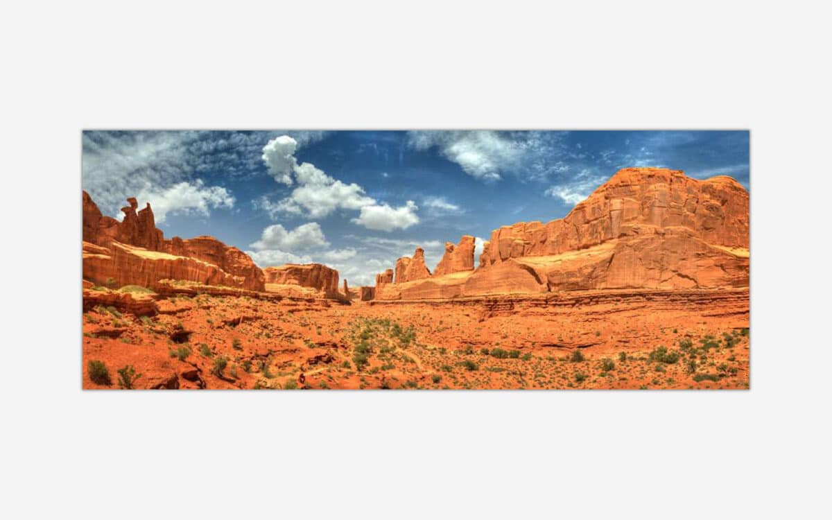 A panoramic image of a desert landscape featuring red rock formations under a blue sky with clouds at Arches National Park.