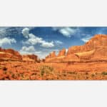 A panoramic image of a desert landscape featuring red rock formations under a blue sky with clouds at Arches National Park.
