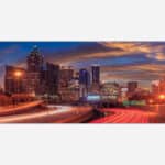 A panoramic view of a city skyline at dusk with illuminated buildings and light trails from moving vehicles on the highway.