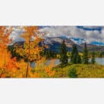 A panoramic autumn landscape with vibrant orange and yellow foliage, a serene lake, pine trees, and a mountain background under a cloudy sky.