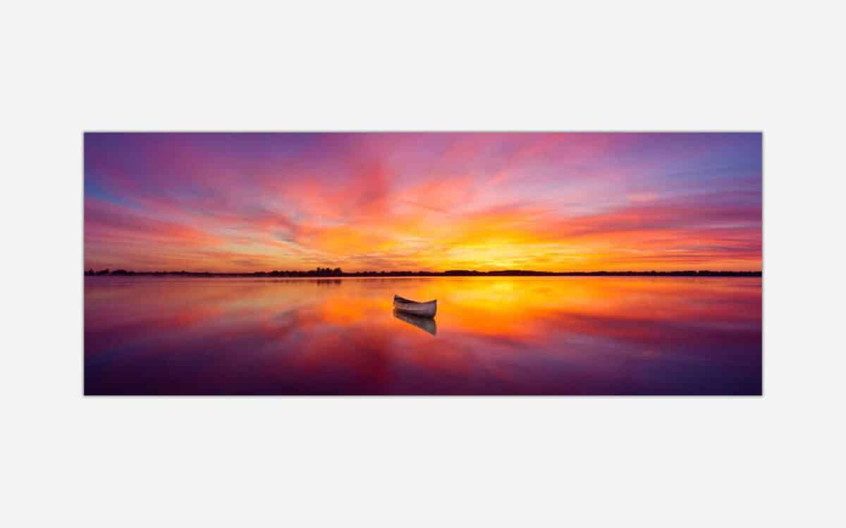 A panoramic photograph of a lone boat on a still lake with a vibrant and colorful sunset reflecting on the water.