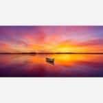 A panoramic photograph of a lone boat on a still lake with a vibrant and colorful sunset reflecting on the water.