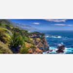 A panoramic view of a coastal landscape with a rocky shoreline, blue ocean, greenery, and a clear sky.