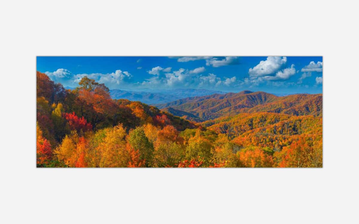 Panoramic autumn landscape with a vibrant display of fall colors on forested mountains under a blue sky with clouds.