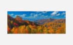 Panoramic autumn landscape with a vibrant display of fall colors on forested mountains under a blue sky with clouds.