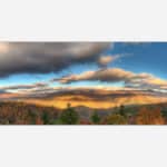 A panoramic painting of a mountain landscape during autumn with colorful trees and sunset-lit clouds in the sky.