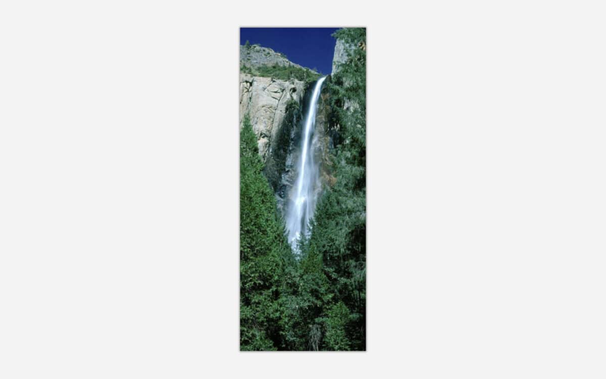 A vertical panoramic photograph depicting a tall waterfall cascading down a rocky cliff surrounded by lush green forest.