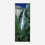 A vertical panoramic photograph depicting a tall waterfall cascading down a rocky cliff surrounded by lush green forest.