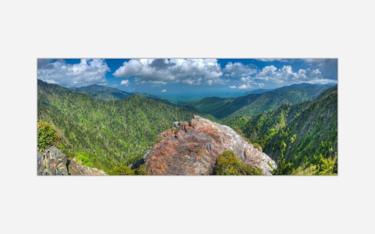 A wide panoramic photograph of a scenic mountain landscape with lush greenery and a vibrant blue sky with clouds.