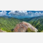 A wide panoramic photograph of a scenic mountain landscape with lush greenery and a vibrant blue sky with clouds.