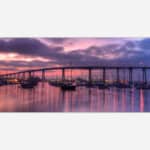 A panoramic photograph of a marina with boats and a bridge in the background under a purple sunset sky.