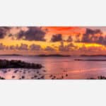 A vibrant painting of a sunset over a marina filled with boats, with a backdrop of islands and a colorful sky reflected in the water.