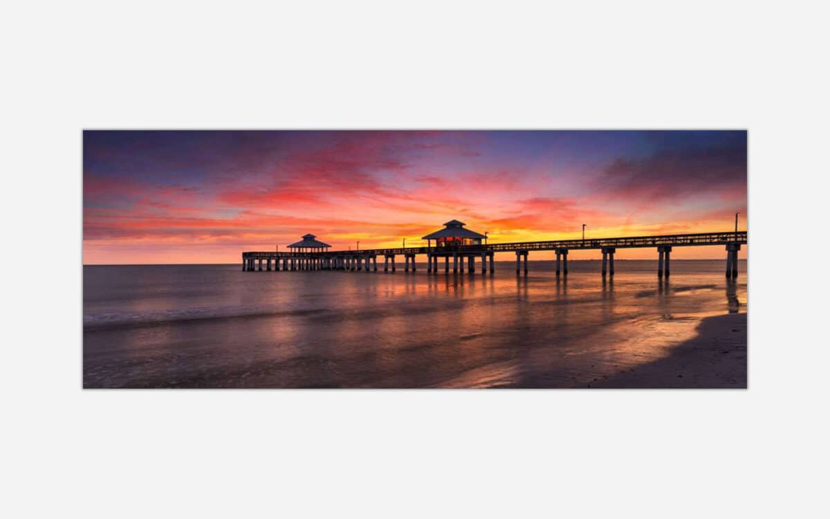 A photo of a pier extending into the ocean with a vibrant sunset sky reflected on the water's surface.