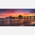A photo of a pier extending into the ocean with a vibrant sunset sky reflected on the water's surface.