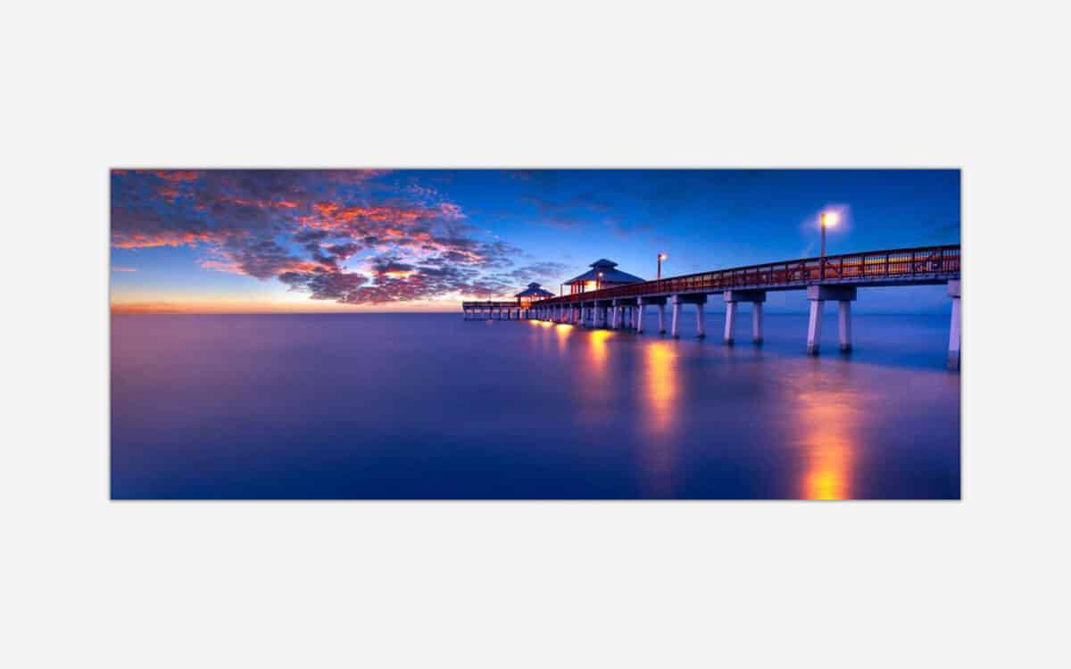 A peaceful seascape photograph featuring a pier extending into the ocean with a stunning sunset sky of blue and orange hues, and reflections of the pier's lights in the water.