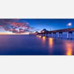 A peaceful seascape photograph featuring a pier extending into the ocean with a stunning sunset sky of blue and orange hues, and reflections of the pier's lights in the water.