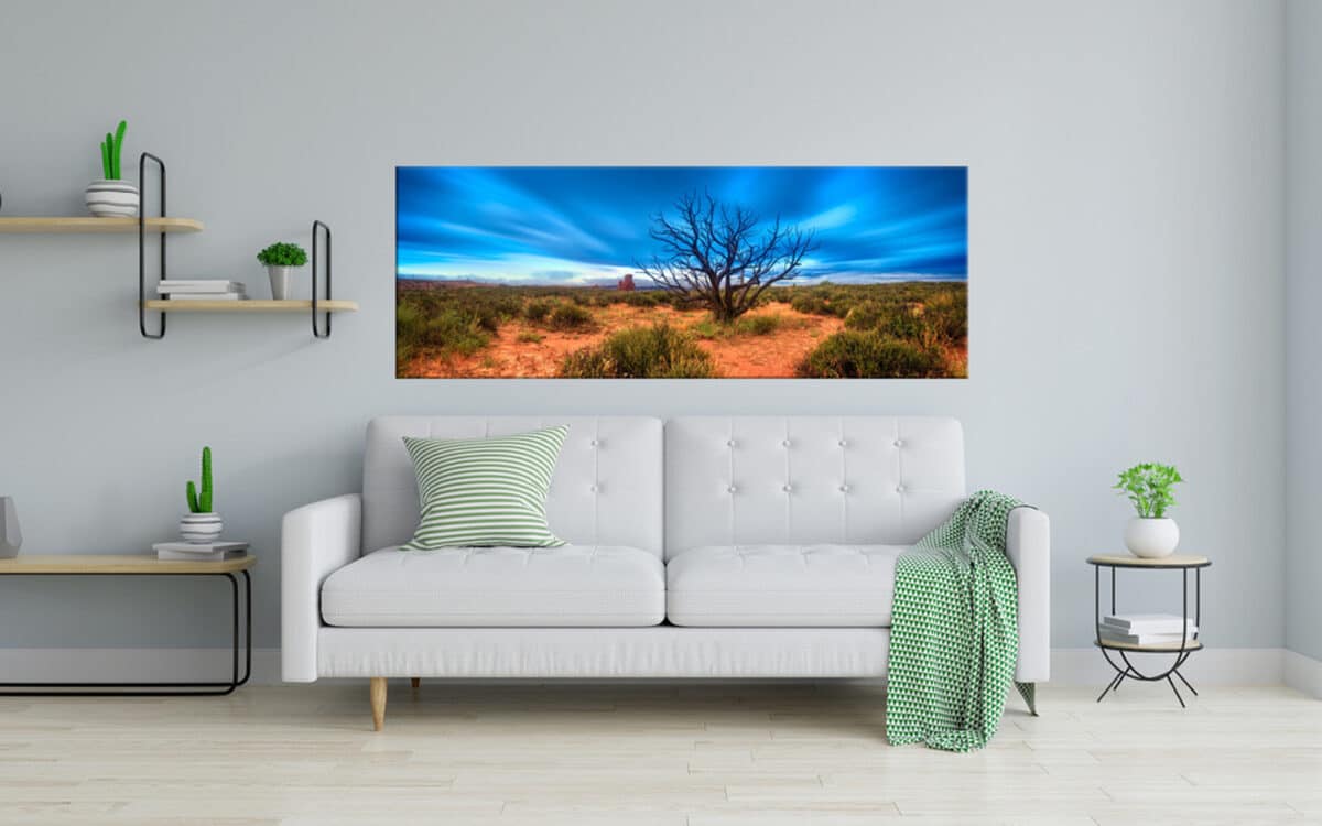 A panoramic landscape canvas depicting a vibrant blue sky and red desert, mounted above a white sofa in a modern living room setup.