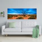 A panoramic landscape canvas depicting a vibrant blue sky and red desert, mounted above a white sofa in a modern living room setup.