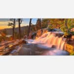 An artful photograph of a waterfall with the sun rising in the background, illuminating the trees and water with a warm golden light, suitable for decorative wall art.