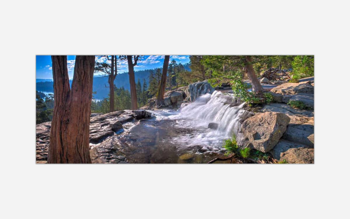 A panoramic photograph of a small waterfall with cascading water through a rocky forest stream, framed by trees against a bright blue sky.