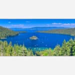 Panoramic view of the blue waters of Lake Tahoe with surrounding green forests and mountain ranges under a clear sky.