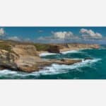 A seascape wall art depicting turquoise ocean waters crashing against rugged cliffs under a cloudy blue sky.