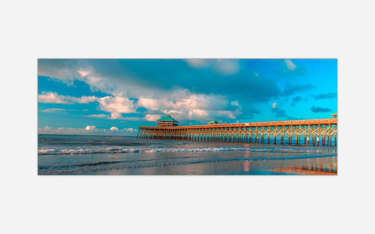 A vibrant photograph of a wooden pier extending into the ocean with waves lapping the shore under a blue sky with scattered clouds.