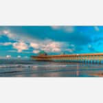 A vibrant photograph of a wooden pier extending into the ocean with waves lapping the shore under a blue sky with scattered clouds.