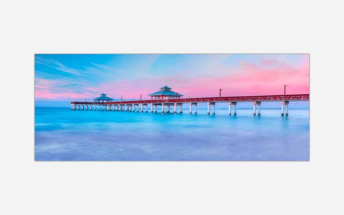 A panoramic photograph of a serene seascape with a pier extending into calm blue waters under a pastel pink and blue sunset sky.