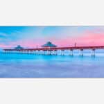 A panoramic photograph of a serene seascape with a pier extending into calm blue waters under a pastel pink and blue sunset sky.