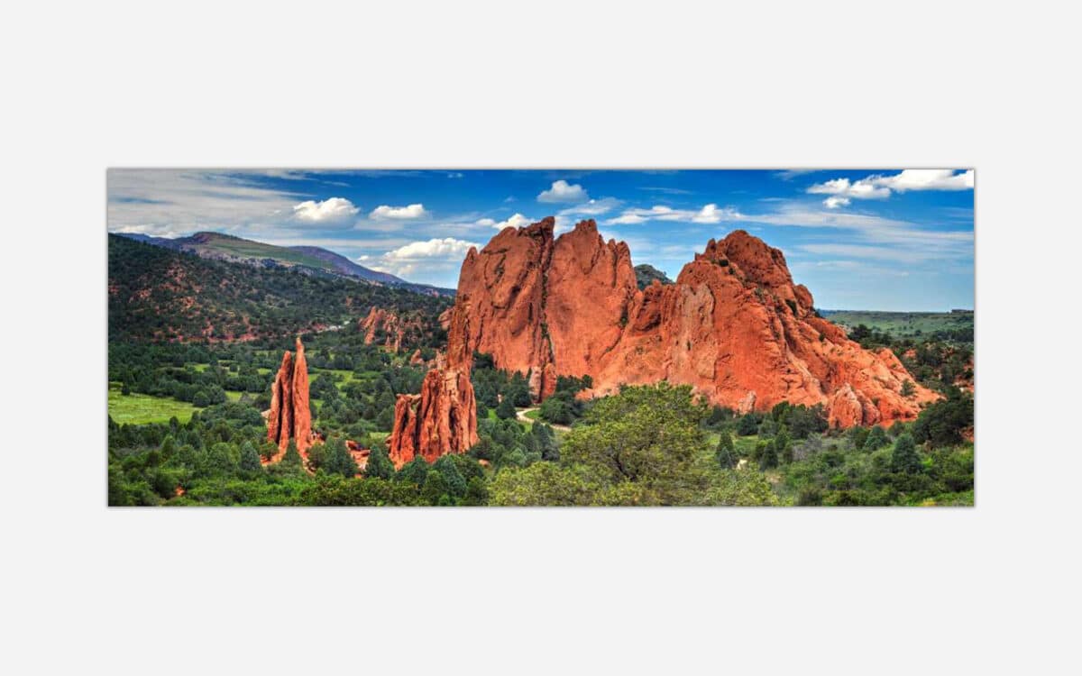 A panoramic photograph of the Garden of the Gods with red rock formations, green vegetation, and a blue sky with clouds.