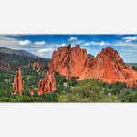 A panoramic photograph of the Garden of the Gods with red rock formations, green vegetation, and a blue sky with clouds.