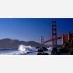 Golden Gate Bridge with ocean waves crashing on the shore and a clear blue sky in the background.