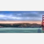 A panoramic photograph of the Golden Gate Bridge at sunset with colorful skies and calm sea in the foreground.