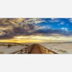 A panoramic image of a wooden walkway leading to a sunset on the beach with vibrant clouds and dune grass.