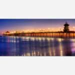 A serene sunset seascape showing a pier extending into the ocean with reflections of lights on the water.