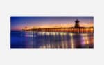 A serene sunset seascape showing a pier extending into the ocean with reflections of lights on the water.