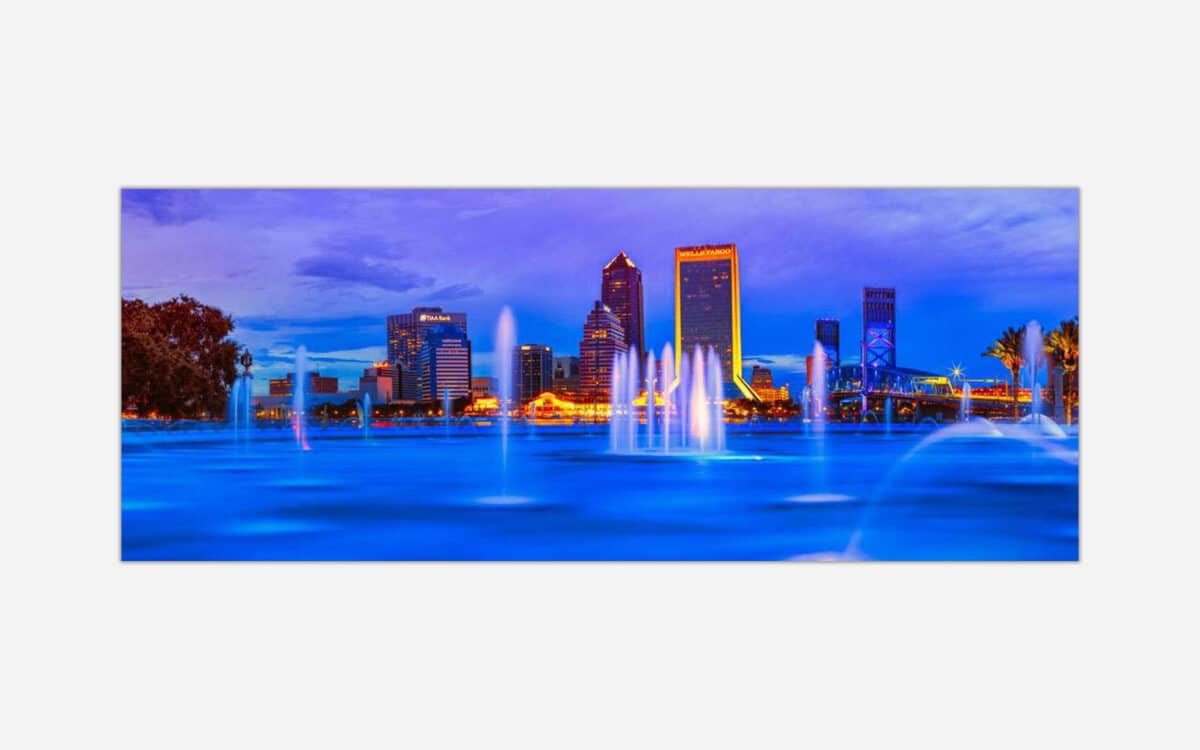 Long exposure photograph of a city skyline at dusk with illuminated buildings and fountain lights, with smooth water foreground due to long exposure technique.