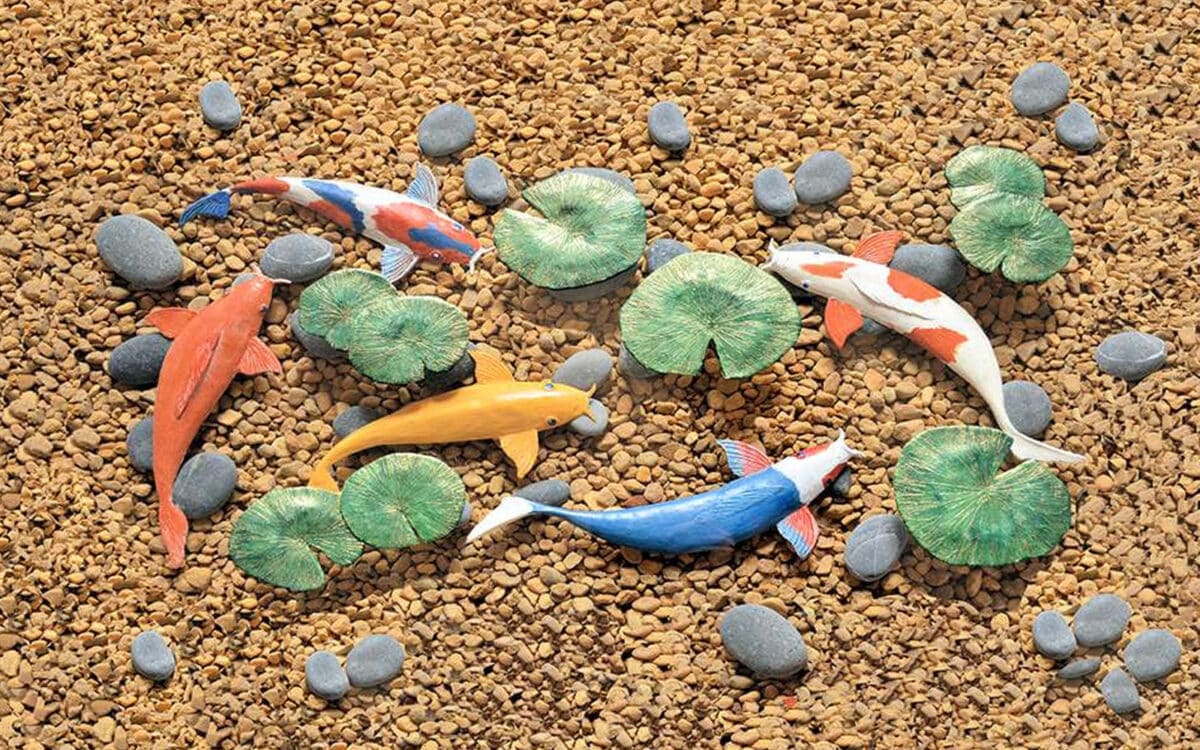 Artistic depiction of a koi pond with colorful painted koi fish, water lily pads, and pebbles on a gravel background.