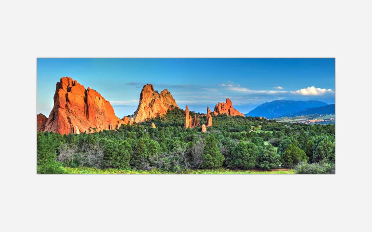 A panoramic landscape photo of majestic orange rock formations against a blue sky with lush greenery in the foreground.