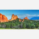 A panoramic landscape photo of majestic orange rock formations against a blue sky with lush greenery in the foreground.