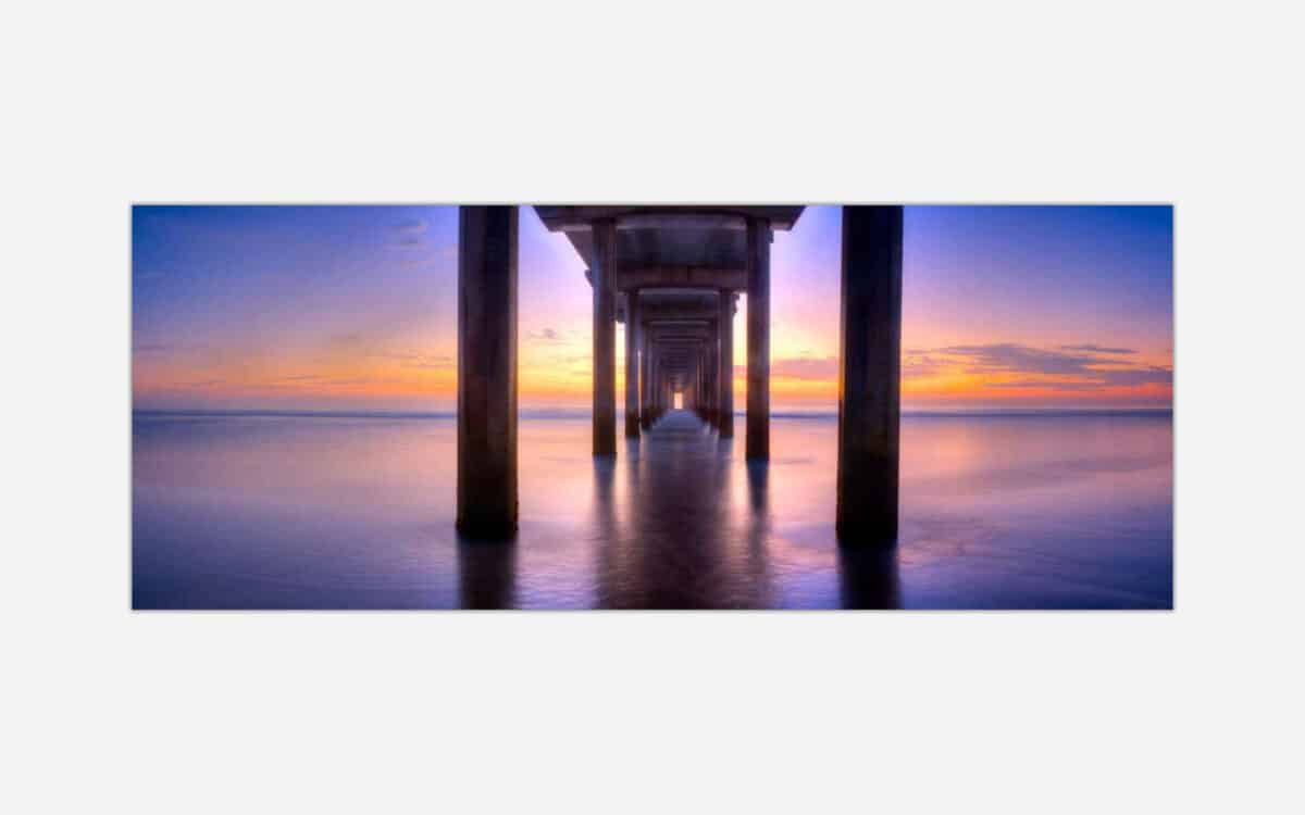 Underneath a pier with calm waters and a vibrant sunset sky in the background, creating a serene and tranquil atmosphere.