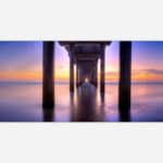 Underneath a pier with calm waters and a vibrant sunset sky in the background, creating a serene and tranquil atmosphere.