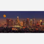 A panoramic view of an illuminated city skyline at night with a full moon in the sky.