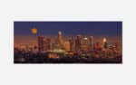 A panoramic view of an illuminated city skyline at night with a full moon in the sky.