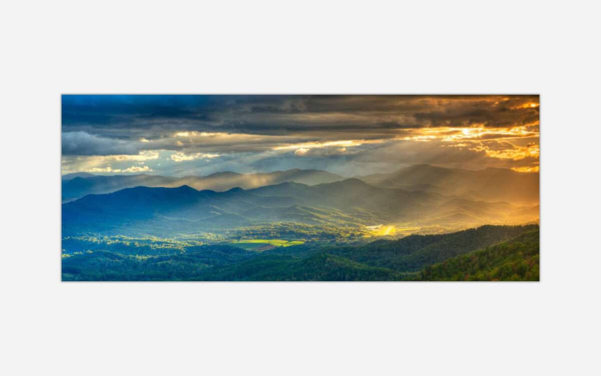 A panoramic landscape photograph of a mountain range during sunset with sunbeams breaking through clouds, casting a golden light over the valleys.