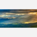 A panoramic landscape photograph of a mountain range during sunset with sunbeams breaking through clouds, casting a golden light over the valleys.