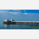 A photograph of a long wooden pier extending into the calm blue ocean under a clear sky.