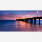An image of a pier extending into the ocean with a vibrant sunset in the background and light reflections on water.