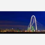 A panoramic view of the illuminated Dallas skyline at night with the prominent Margaret Hunt Hill Bridge in the foreground.